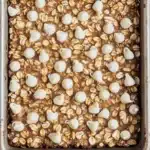 Sugar cookie overnight oats - the image shows a rectangular baking tray filled with granola bars. the bars are golden brown in color and appear to be freshly baked. on top of the granola, there are small white chocolate hearts scattered across the surface. the tray is lined with parchment paper and is placed on a white wooden surface.