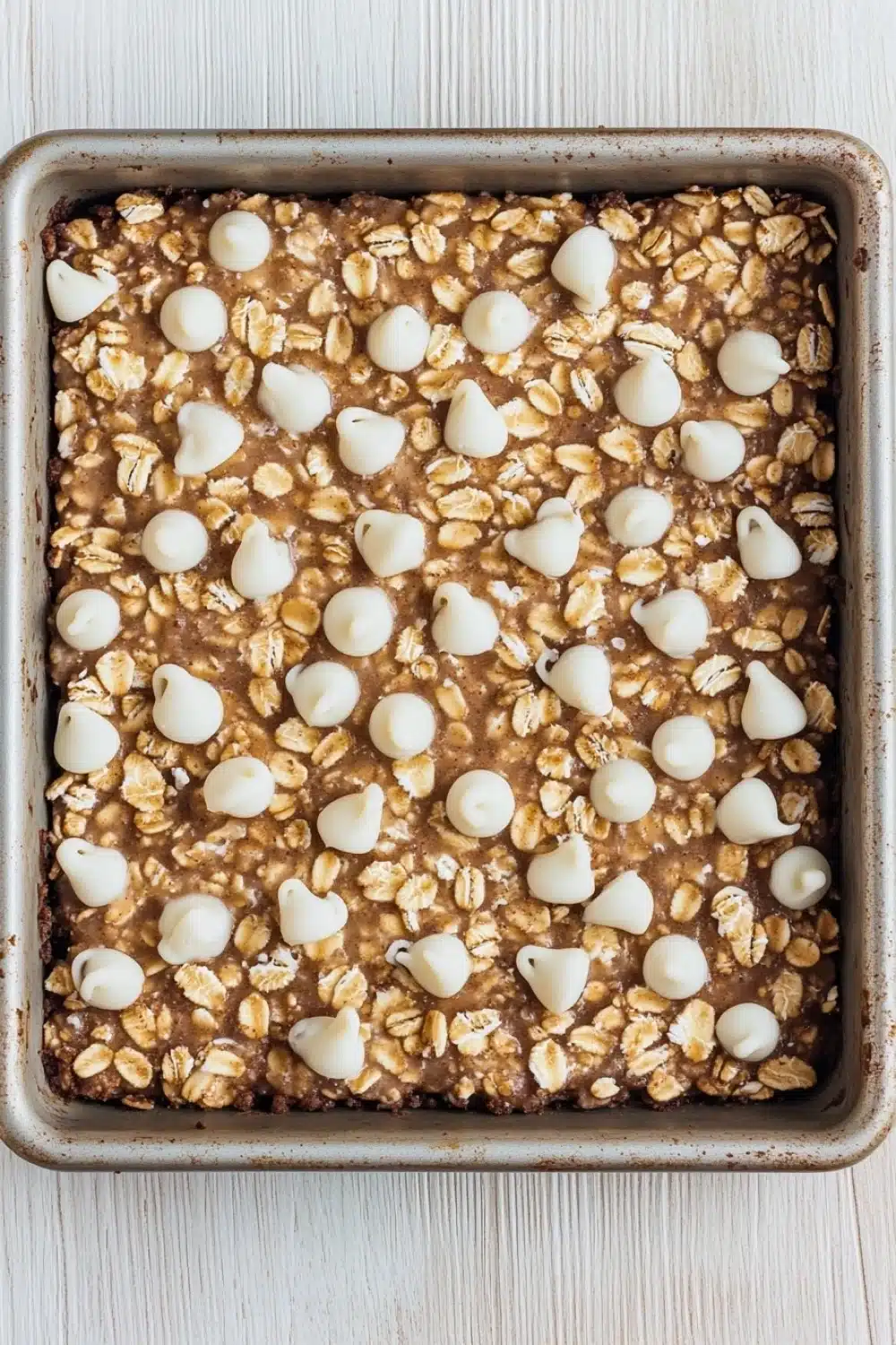 Sugar cookie overnight oats - the image shows a rectangular baking tray filled with granola bars. the bars are golden brown in color and appear to be freshly baked. on top of the granola, there are small white chocolate hearts scattered across the surface. the tray is lined with parchment paper and is placed on a white wooden surface.