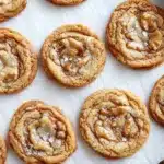Thin and crispy cookie - the image shows a tray of freshly baked cookies on a white parchment paper. the cookies are round and golden brown in color, with a swirl pattern on top. they are arranged in a single layer on the tray, with some overlapping each other. some of the cookies have a light dusting of powdered sugar on top, while others have a darker brown color. the tray is lined with parchment paper, and there is a metal tray on the right side of the image. the background is blurred, but it appears to be a kitchen countertop.