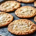 Thin and crispy cookie - the image shows a baking tray with freshly baked cookies on it. the cookies are round and golden brown in color, with a crumbly texture on top. they are arranged in a single layer on the tray, with some overlapping each other. the tray is lined with parchment paper, and there is a metal spatula resting on the side. the background is blurred, but it appears to be a kitchen countertop.