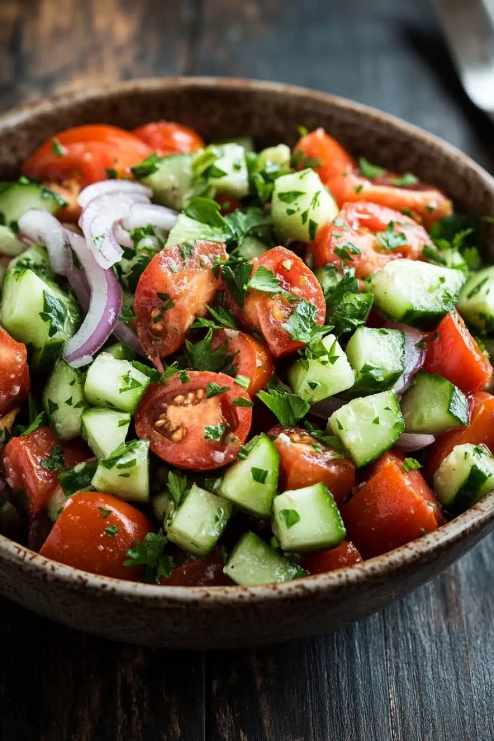 Tomato cucumber and onion salad - the image is a close-up of a bowl of salad. the salad is made up of diced tomatoes, cucumbers, and red onions. the tomatoes are cut into small cubes and are arranged in a circular pattern. the cucumbers are cut in half and are scattered throughout the salad, with some of them overlapping each other. there are also some chopped herbs, such as cilantro, scattered throughout. the bowl is made of dark wood and is sitting on a wooden table. the background is blurred, but it appears to be a rustic kitchen countertop.