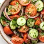 Tomato cucumber and onion salad - the image is a close-up of a bowl of salad. the bowl is made of ceramic and has a light brown color. the salad is made up of sliced tomatoes, cucumbers, red onions, and chopped herbs. the tomatoes are cut into thin slices and are arranged in a circular pattern. the cucumbers are sliced into thin strips and are scattered throughout the salad, with some of them overlapping each other. the herbs are finely chopped and add a pop of green color to the dish. the background is white, making the colors of the salad stand out.