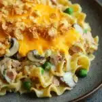 Tuna casserole classic - the image shows a plate of pasta dish. the pasta appears to be fettuccine alfredo, with a layer of melted cheese on top. the cheese is golden brown and bubbly, and it is drizzled over the top with crumbled breadcrumbs. there are also chunks of ham and mushrooms scattered throughout the dish, as well as some green peas. the dish is served on a dark brown plate with a dark background.