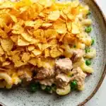 Tuna casserole with potato chips - the image is a close-up of a plate of macaroni and cheese. the plate is round and has a light brown rim with a speckled pattern. on top of the macaronis, there is a layer of shredded cheese, which is a bright yellow color. the cheese appears to be melted and bubbly, and there are small pieces of chicken and peas scattered throughout the dish. the dish is garnished with a sprinkle of green peas. the background is a white wooden table.