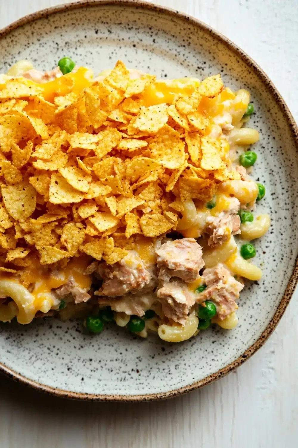 Tuna casserole with potato chips - the image is a close-up of a plate of macaroni and cheese. the plate is round and has a light brown rim with a speckled pattern. on top of the macaronis, there is a layer of shredded cheese, which is a bright yellow color. the cheese appears to be melted and bubbly, and there are small pieces of chicken and peas scattered throughout the dish. the dish is garnished with a sprinkle of green peas. the background is a white wooden table.