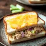 Tuna melt sandwiches - the image shows a close-up of a sandwich on a plate. the sandwich is made with two slices of white bread and is cut in half, revealing the filling inside. the filling appears to be a mixture of chicken, celery, and onions. the cheese is melted and oozing out of the sides of the sandwich. the plate is on a wooden table with a few celery sticks scattered around it. in the background, there is another plate with more sandwiches on it. the overall mood of the image is rustic and appetizing.