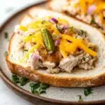 Tuna melt with relish - the image shows a plate with two slices of bread on it. the bread appears to be toasted and has a golden brown crust. on top of the bread, there is a mixture of shredded chicken, diced red onions, and yellow cheese. there is also a pickle on top. the plate is garnished with chopped parsley. the background is a white marble countertop.