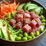 Tuna poke bowl spicy - the image is a close-up of a bowl of food. the bowl is made of ceramic and is placed on a dark grey countertop. the food in the bowl appears to be a salad with various ingredients. on top of the salad, there are sliced cucumbers, sliced carrots, sliced avocado, and edamame. the salad is topped with sesame seeds and green onions. the colors of the ingredients are vibrant and the dish looks fresh and appetizing.