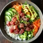 Tuna poke bowl spicy - the image is a close-up of a bowl of food on a wooden table. the bowl is made of dark grey ceramic and is filled with a bed of white rice. on top of the rice, there are sliced cucumbers, sliced carrots, sliced avocado, and sliced green onions. the vegetables are arranged in a colorful and appetizing manner, with some of them overlapping each other. there are also some sesame seeds sprinkled on top, adding a pop of color to the dish. the overall appearance of the dish is fresh and vibrant.