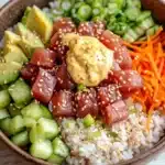 Tuna poke bowl spicy mayo - the image is a close-up of a bowl of food. the bowl is made of dark brown ceramic and is placed on a white wooden table. the food in the bowl appears to be a salad with a variety of ingredients. on top of the salad, there are sliced avocado, sliced cucumbers, sliced carrots, and sliced green onions. there is also a dollop of yellow sauce on top. the salad is topped with sesame seeds and sesame seed. the colors of the ingredients are vibrant and the dish looks fresh and appetizing.