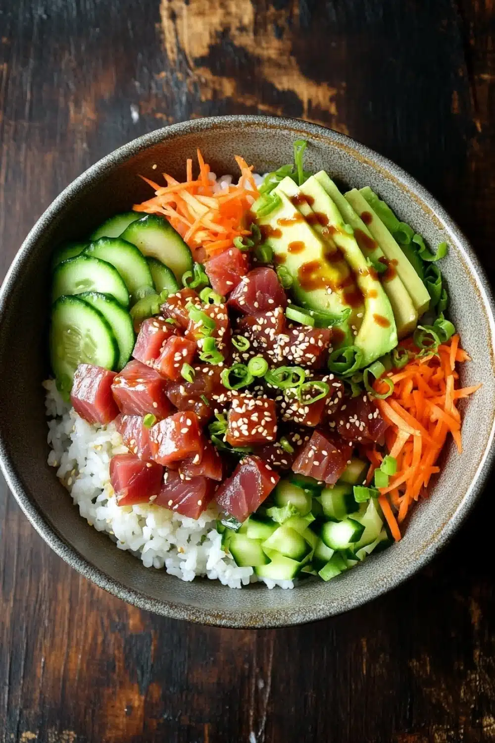 Tuna poke bowl spicy - the image is a close-up of a bowl of food on a wooden table. the bowl is made of dark grey ceramic and is filled with a bed of white rice. on top of the rice, there are sliced cucumbers, sliced carrots, sliced avocado, and sliced green onions. the vegetables are arranged in a colorful and appetizing manner, with some of them overlapping each other. there are also some sesame seeds sprinkled on top, adding a pop of color to the dish. the overall appearance of the dish is fresh and vibrant.