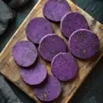 Ube shortbread cookie - the image shows a wooden cutting board with six round, purple-colored slices arranged in a circular pattern on it. the slices appear to be freshly cut and have a glossy texture. the cutting board is placed on a black slate surface, and there is a sprig of thyme on the right side of the image. a gray cloth napkin is visible on the left side. the overall aesthetic is rustic and minimalistic.