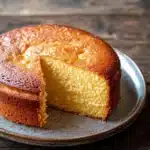 Vanilla cake scratch - the image is of a round cake on a silver plate. the cake appears to be freshly baked and has a golden brown crust on top. a slice of the cake is cut out, revealing the soft, fluffy interior. the plate is placed on a wooden table with a rustic, weathered look. the background is blurred, making the cake the focal point of the image.
