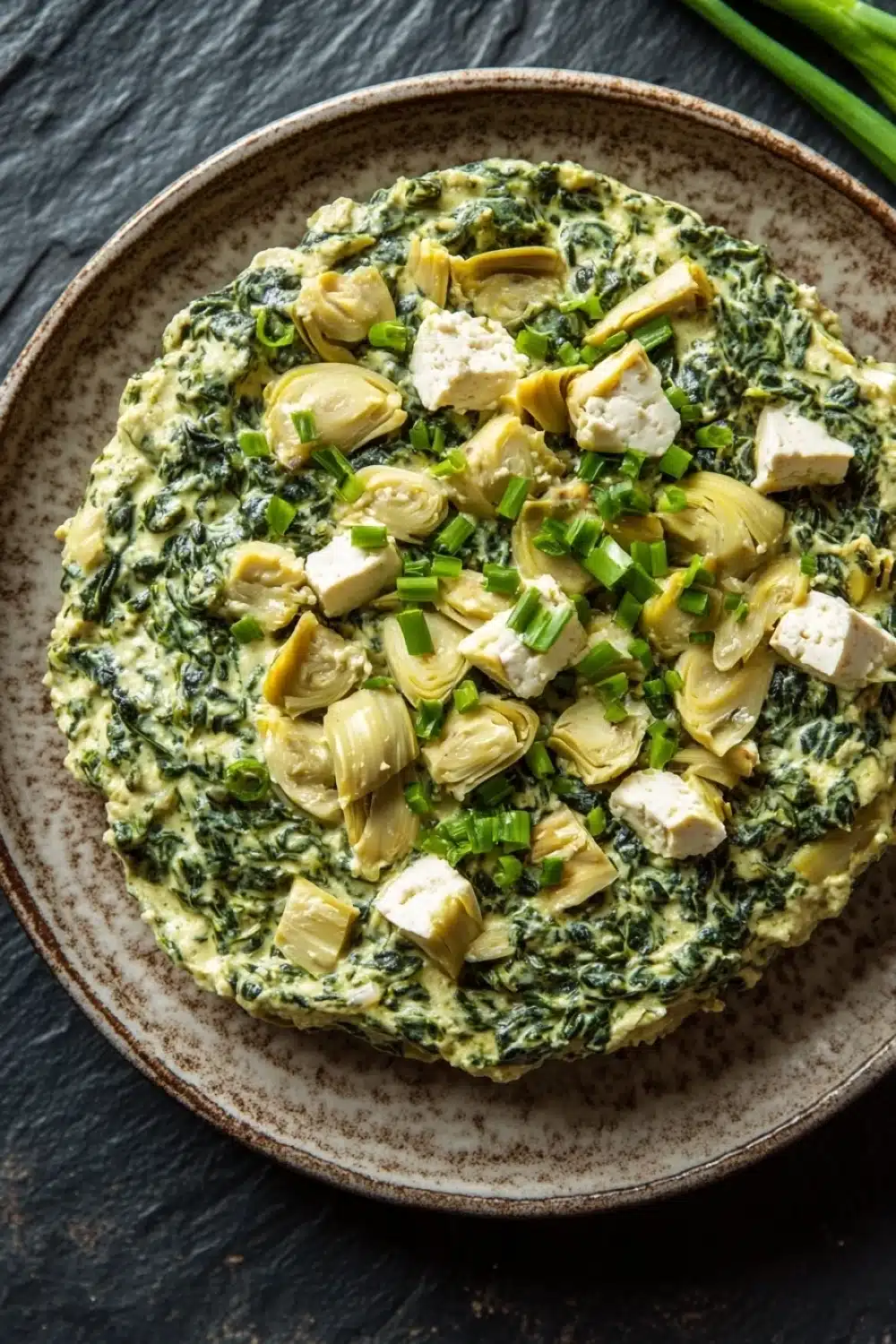 Vegan spinach artichoke dip with tofu - the image is a close-up of an omelette in a brown ceramic bowl. the omelet is made with artichokes, spinach, and feta cheese, and is garnished with chopped green onions. the bowl is sitting on a dark grey textured surface, and there are a few sprigs of fresh green onions scattered around the bowl.