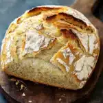 Vegan yeast bread - the image shows a freshly baked loaf of bread on a wooden cutting board. the bread appears to be freshly baked, with a golden brown crust and a soft, fluffy interior. the top of the bread is covered in a layer of melted cheese, which is visible on the surface. the cutting board is placed on a dark, textured surface, possibly a table or countertop. the background is blurred, making the bread the focal point of the image.