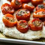 Whipped feta dip roasted tomatoes - the image shows a tray of roasted cherry tomatoes on a baking sheet. the tomatoes are arranged in a circular pattern on the tray, with some overlapping each other. they are bright red in color and appear to be seasoned with herbs and spices. on top of the tomatoes, there is a layer of creamy white sauce, which looks like feta cheese. the tray is lined with parchment paper, and the background is blurred, making the tomatoes the focal point of the image.