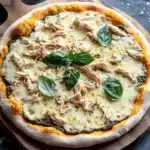 White chicken pesto pizza - the image shows a round pizza on a wooden cutting board. the pizza is topped with melted cheese and chunks of chicken. there are also fresh basil leaves scattered around the board, adding a pop of green color to the dish. in the background, there is a small clay pot and a few scattered salt. the cutting board is placed on a dark grey countertop.