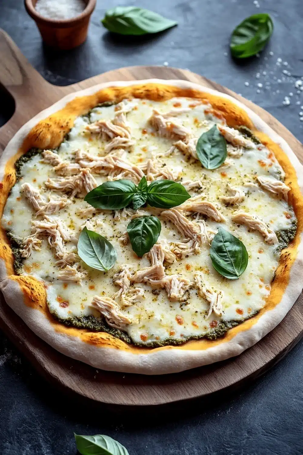 White chicken pesto pizza - the image shows a round pizza on a wooden cutting board. the pizza is topped with melted cheese and chunks of chicken. there are also fresh basil leaves scattered around the board, adding a pop of green color to the dish. in the background, there is a small clay pot and a few scattered salt. the cutting board is placed on a dark grey countertop.