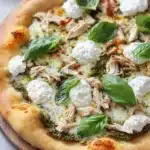 White chicken pesto pizza - the image is of a freshly baked pizza on a wooden cutting board. the pizza has a golden brown crust and is topped with shredded chicken, mozzarella cheese, and fresh basil leaves. the cheese is melted and bubbly, and the basil leaves are scattered around the pizza. the background is a white countertop with a few more basil leaves scattered around.