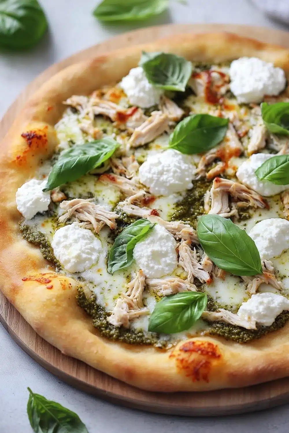 White chicken pesto pizza - the image is of a freshly baked pizza on a wooden cutting board. the pizza has a golden brown crust and is topped with shredded chicken, mozzarella cheese, and fresh basil leaves. the cheese is melted and bubbly, and the basil leaves are scattered around the pizza. the background is a white countertop with a few more basil leaves scattered around.