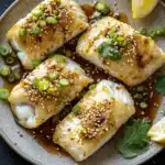 White fish asian - the image shows a plate of four pieces of fish fillets covered in sesame seeds and green onions. the fillets are arranged in a circular pattern on a dark grey plate. there are two lemon wedges on the side of the plate and a sprig of cilantro on the right side. the plate is garnished with a few sprigs of parsley. the overall presentation of the dish is elegant and appetizing.