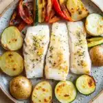 White fish dinner - the image shows a plate of food on a wooden table. on the plate, there are three pieces of fish fillets arranged in a circular pattern. the fillets are golden brown and appear to be seasoned with herbs and spices. next to the fillets, there is a serving of roasted vegetables, including zucchini, red bell peppers, and yellow squash. the vegetables are arranged in an appetizing manner, with some of them overlapping each other. the plate is garnished with a sprig of parsley. the background is blurred, but it appears to be a kitchen countertop.