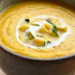 Zucchini and butternut squash soup - the image is a close-up of a bowl of orange-colored soup. the soup appears to be thick and creamy, with a smooth texture. on top of the soup, there are small pieces of sliced zucchini arranged in a circular pattern. the bowl is made of ceramic and has a dark brown color. the background is blurred, making the soup the focal point of the image.