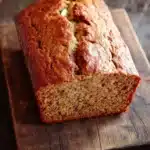 Zucchini banana bread - the image shows a freshly baked loaf of bread on a wooden cutting board. the bread appears to be golden brown in color and has a slightly crumbly texture. it is cut in half, revealing the soft, fluffy interior. the cutting board is placed on a dark wooden table with a rustic, weathered look. the background is blurred, making the bread the focal point of the image.