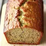 Zucchini banana bread - the image shows a freshly baked loaf of bread on a wooden cutting board. the bread appears to be golden brown in color and has a slightly crumbly texture. it is cut in half, revealing the soft, fluffy interior. the edges of the bread are slightly frayed, indicating that it has been freshly baked. the cutting board is placed on a gray wooden table with a rustic background.