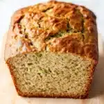 Zucchini bread extra moist - the image shows a freshly baked loaf of bread on a wooden cutting board. the bread appears to be golden brown in color and has a crumbly texture. it is topped with zucchini seeds, which are visible in the center of the loaf. the loaf is sitting on a white countertop with a blurred background.