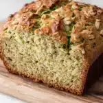 Zucchini bread extra moist - the image shows a freshly baked loaf of bread on a wooden cutting board. the bread appears to be a zucchini bread with a golden brown crust and a soft, fluffy interior. the top of the bread is covered in chopped nuts, giving it a crunchy texture. the cutting board is placed on a white marble countertop.