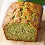 Zucchini bread gluten free dairy free - the image shows a freshly baked loaf of zucchini bread on a wooden cutting board. the bread is golden brown in color and has a crumbly texture. it appears to be moist and fluffy, with visible zucchinis and herbs scattered throughout. in the background, there is a whole zucchi, which is a bright green color and is slightly blurred. a white cloth napkin is visible on the right side of the image.