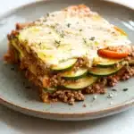 Zucchini tomato ground beef casserole - the image shows a slice of lasagna on a gray plate. the lasagna is made with layers of ground beef, zucchini, and tomato sauce. the top layer is covered in a layer of melted cheese and sprinkled with chopped herbs. the plate is sitting on a white countertop. a fork is resting on the side of the plate.