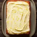 9 x 13 lemon cake - the image shows a rectangular baking tray with a freshly baked cake on it. the cake appears to be a carrot cake with a golden brown crust and a creamy white frosting on top. the frosting is spread evenly across the top of the cake, creating a swirl pattern. there are small yellow sprinkles scattered throughout the frosting, adding a pop of color to the dish. the tray is placed on a wooden table, giving the image a rustic feel.