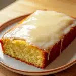 9 x 13 lemon cake - the image shows a slice of cake on a white plate. the cake appears to be freshly baked and has a golden brown crust on top. the top of the cake is covered in a thick layer of white icing, which is drizzled over the cake. the plate is sitting on a wooden cutting board. the background is blurred, but it seems to be a kitchen countertop.