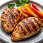 Air fryer chicken breast and peppers - the image shows two grilled chicken breasts on a gray plate. the chicken breasts are golden brown and appear to be seasoned with herbs and spices. on top of the chicken breasts, there are sliced red and yellow bell peppers and a sprig of parsley. the plate is sitting on a white marble countertop.