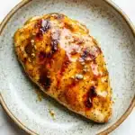 Air fryer chicken breast glazed - the image shows a piece of grilled chicken on a white marble plate. the chicken is golden brown and appears to be seasoned with sesame seeds and herbs. the plate is round and has a light blue rim. the background is blurred, making the chicken the focal point of the image.