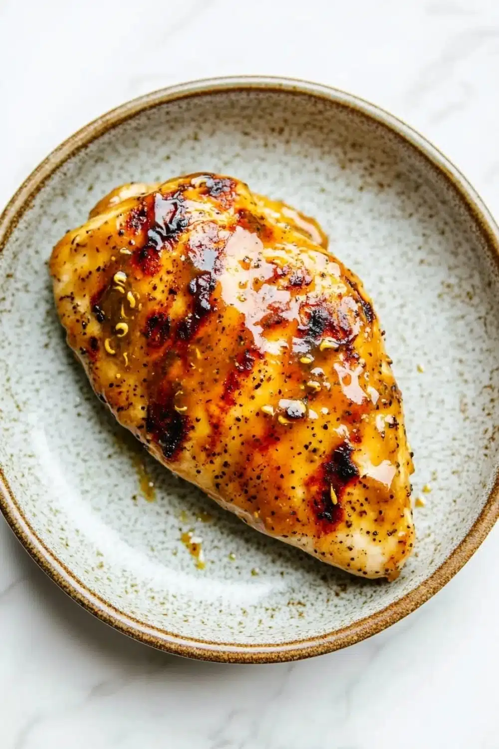 Air fryer chicken breast glazed - the image shows a piece of grilled chicken on a white marble plate. the chicken is golden brown and appears to be seasoned with sesame seeds and herbs. the plate is round and has a light blue rim. the background is blurred, making the chicken the focal point of the image.