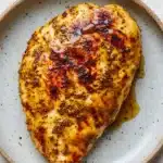 Air fryer chicken breast honey mustard - the image shows a plate with a piece of grilled chicken on it. the chicken is golden brown and appears to be seasoned with herbs and spices. it is covered in a thick layer of melted cheese and has a drizzle of sauce on top. the plate is light blue with a speckled pattern around the edges. the background is a white wooden table.