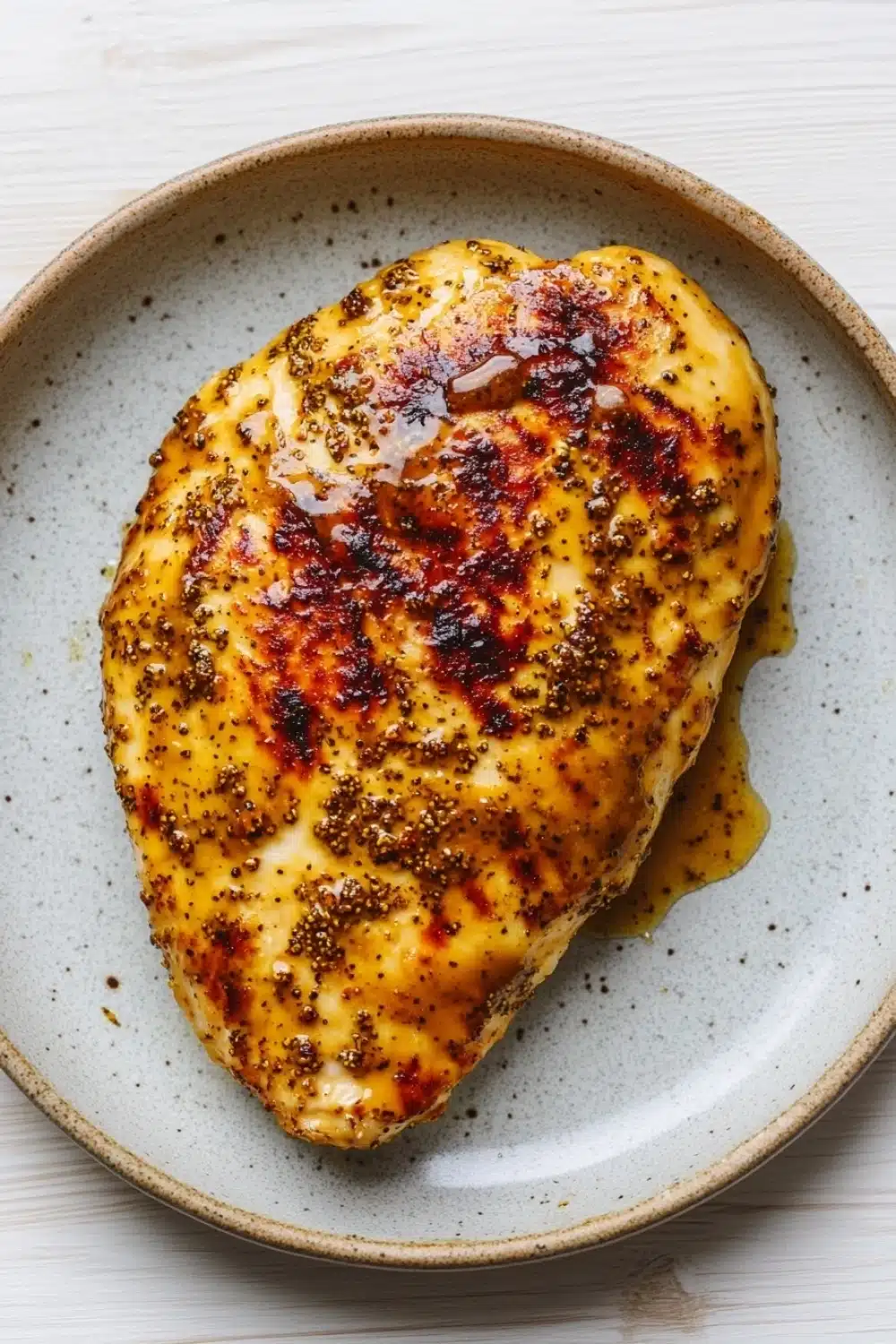 Air fryer chicken breast honey mustard - the image shows a plate with a piece of grilled chicken on it. the chicken is golden brown and appears to be seasoned with herbs and spices. it is covered in a thick layer of melted cheese and has a drizzle of sauce on top. the plate is light blue with a speckled pattern around the edges. the background is a white wooden table.