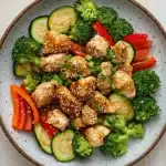 Air fryer chicken teriyaki with vegetables - the image is a close-up of a bowl of asian-style stir-fry. the bowl is made of ceramic and has a light brown color with a speckled pattern. it is filled with a variety of vegetables, including broccoli, zucchini, red bell peppers, and sliced cucumbers. the vegetables are arranged in a colorful and appetizing manner, with some pieces of chicken and sesame seeds sprinkled on top. the chicken is cut into small cubes and appears to be seasoned with herbs and spices. the colors of the vegetables are vibrant and the dish looks fresh and vibrant.