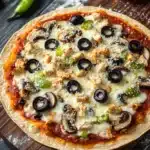 Air fryer deep dish tortilla pizza - the image is of a pizza on a wooden cutting board. the pizza is round and has a golden brown crust. it is topped with melted cheese, sliced mushrooms, green bell peppers, and black olives. there are also two whole garlic cloves and two green chili peppers on the cutting board next to the pizza. the background is a dark wooden table.