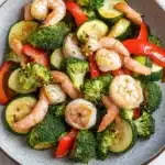 Air fryer shrimp and vegetables - the image is a close-up of a bowl of stir-fry with shrimp and vegetables. the bowl is made of ceramic and has a light blue and white speckled pattern. the vegetables in the bowl include broccoli, zucchini, and red bell peppers. the shrimp are pink and appear to be cooked, while the vegetables are green and yellow. the dish is garnished with a sprinkle of sesame seeds. the background is a white wooden table.