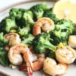 Air fryer shrimp broccoli - the image shows a plate of cooked shrimp and broccoli. the shrimp are pink and appear to be seasoned with herbs and spices. the broccoli is bright green and looks fresh and healthy. there is a slice of lemon on the side of the plate, adding a pop of color to the dish. the plate is white and has a textured surface. the background is blurred, but it appears to be a kitchen countertop.