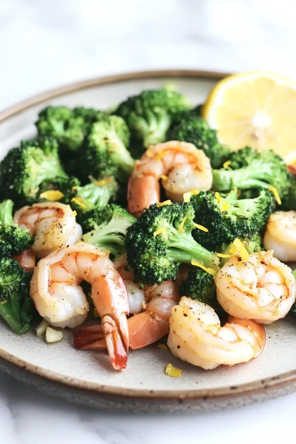 Air fryer shrimp broccoli - the image shows a plate of cooked shrimp and broccoli. the shrimp are pink and appear to be seasoned with herbs and spices. the broccoli is bright green and looks fresh and healthy. there is a slice of lemon on the side of the plate, adding a pop of color to the dish. the plate is white and has a textured surface. the background is blurred, but it appears to be a kitchen countertop.