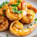 Air fryer shrimp cooking time - the image shows a plate of cooked shrimp on a white marble countertop. the shrimp are golden brown and appear to be seasoned with herbs and spices. they are arranged in a circular pattern on the plate, with some overlapping each other. the plate is garnished with a sprig of fresh parsley. the background is blurred, making the shrimp the focal point of the image.