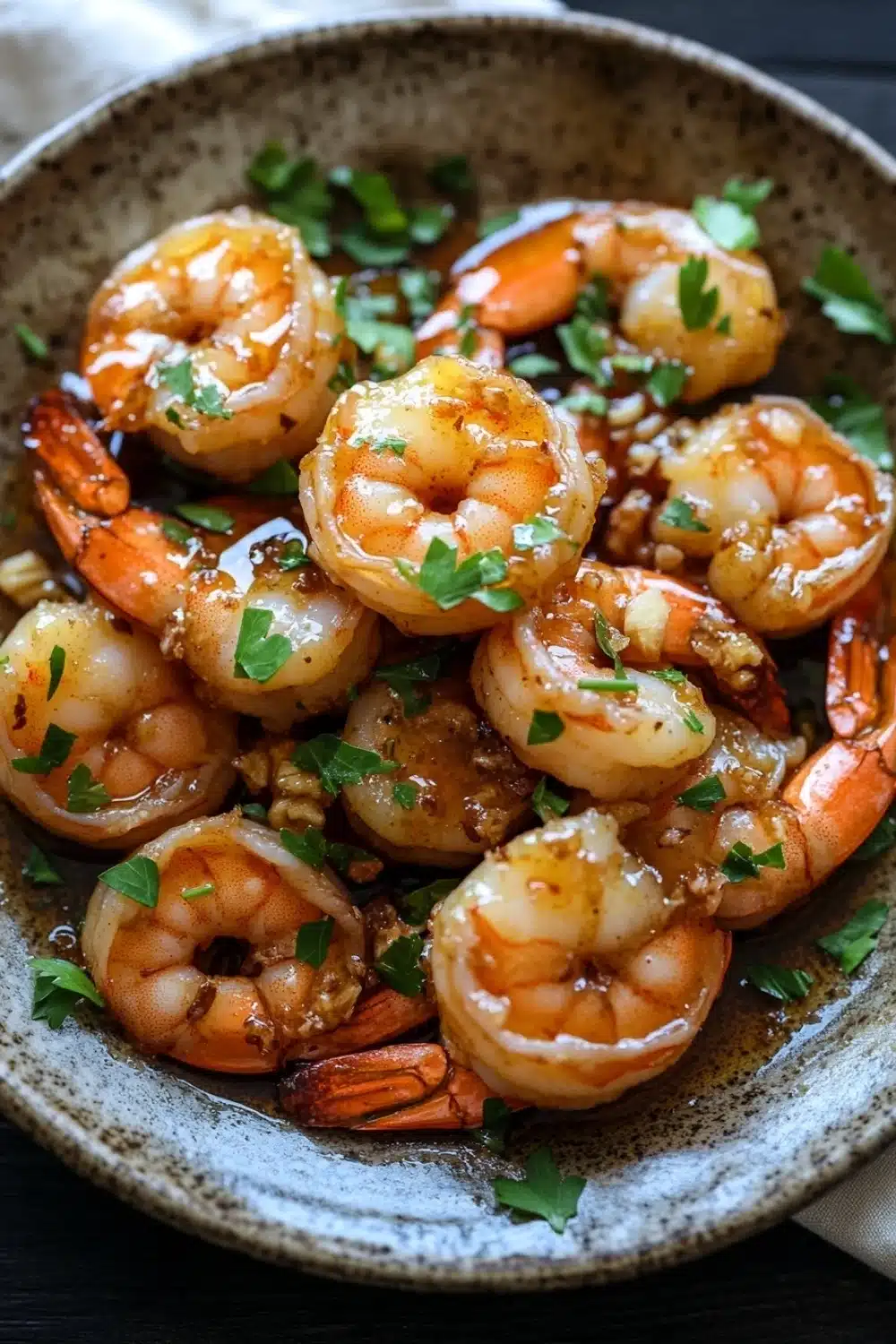 Air fryer shrimp honey garlic - the image shows a plate of cooked shrimp in a brown and white speckled bowl. the shrimp are orange in color and appear to be seasoned with herbs and spices. they are arranged in a circular pattern in the bowl, with some overlapping each other. the dish is garnished with chopped parsley and there is a drizzle of sauce on top. the bowl is sitting on a wooden table with a white napkin in the background.