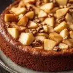 Apple cake from mix boxes - the image is of a freshly baked apple tart on a round cake stand. the tart has a golden brown crust and is topped with sliced apples and pecans. the apples are arranged in a circular pattern on top of the tart, with some overlapping each other. the pecan nuts are scattered throughout the tart and add a crunchy texture to the dish. the cake stand is made of metal and has a blue and white pattern around the edges. the background is a dark wooden table.