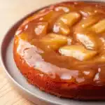 Apple cake glaze - the image is a close-up of a round cake on a gray plate. the cake appears to be freshly baked and has a golden brown crust. on top of the cake, there is a layer of caramel sauce that is drizzled over the top. the caramel sauce is a light brown color and looks smooth and glossy. there are several slices of caramel arranged in a circular pattern on the cake. the plate is sitting on a wooden table.