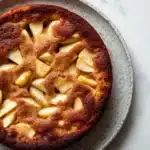 Apple cake no milk - the image is a close-up of a round cake on a white marble countertop. the cake appears to be freshly baked and has a golden brown crust on top. it is topped with sliced apples arranged in a criss-cross pattern, creating a lattice-like design. the apples are cut into small cubes and are arranged evenly across the top of the cake. the plate is round and has small speckles around the edges. the background is blurred, making the cake the focal point of the image.